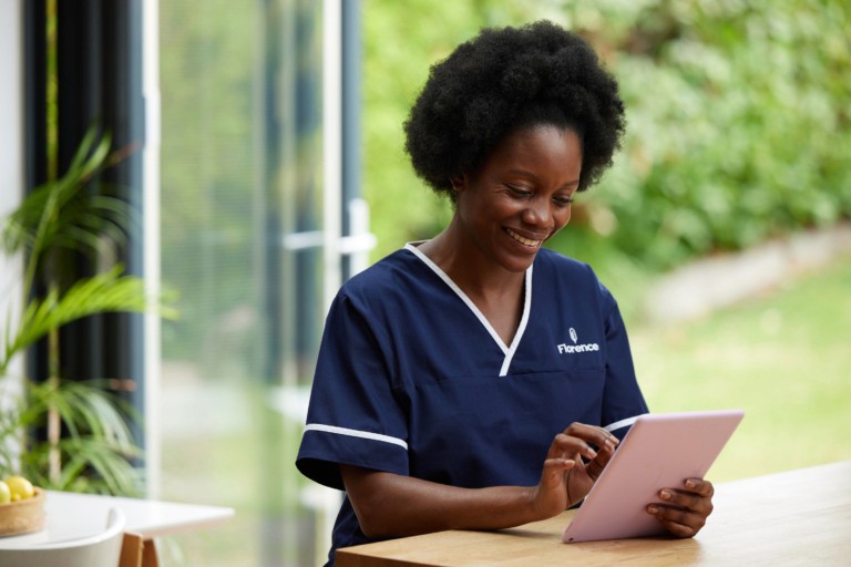 A smiling nurse in a navy blue Florence-branded uniform using a tablet device near a bright window overlooking a garden