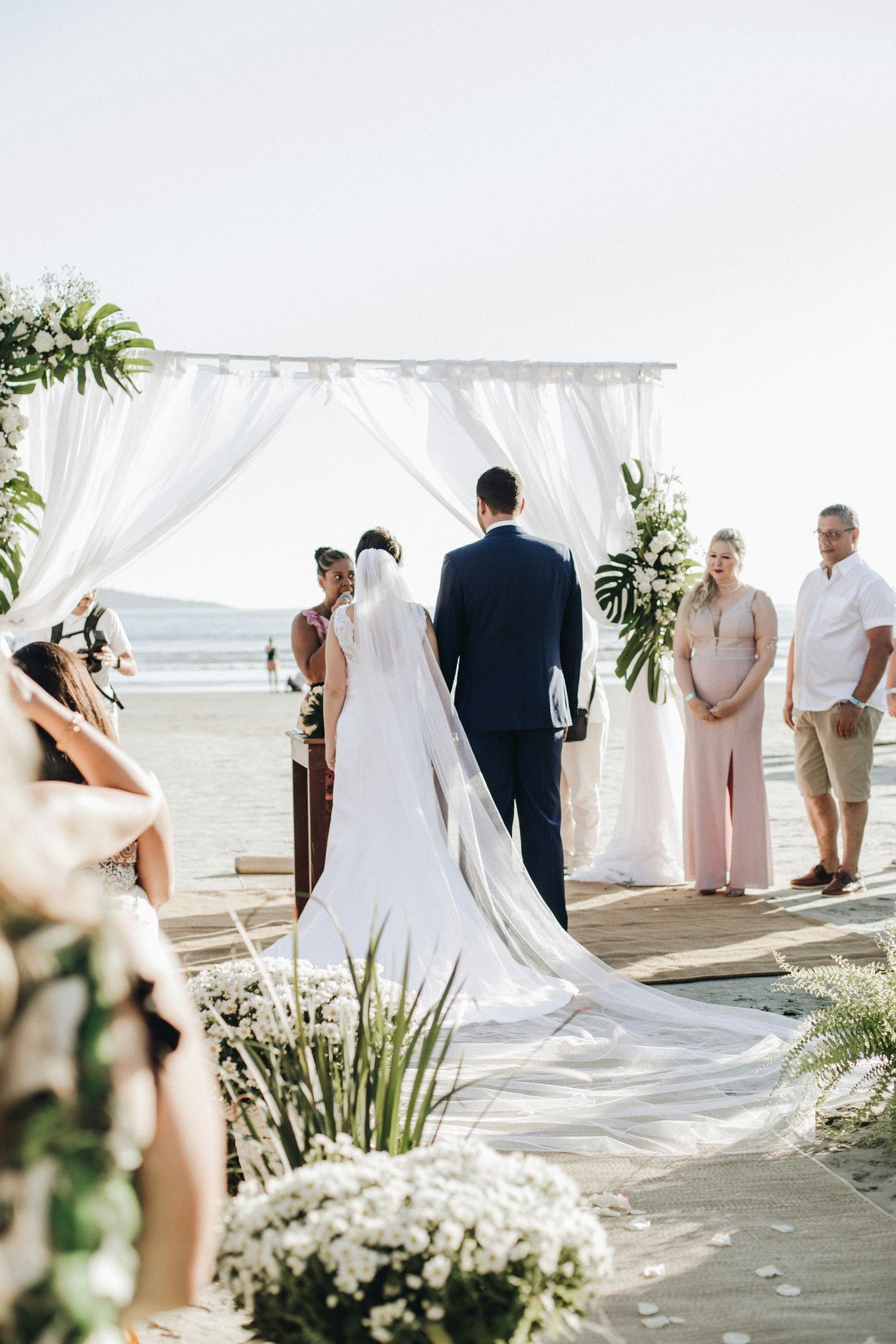 Bride and groom standing at the altar under a white draped canopy during a beachfront wedding ceremony with tropical floral arrangements