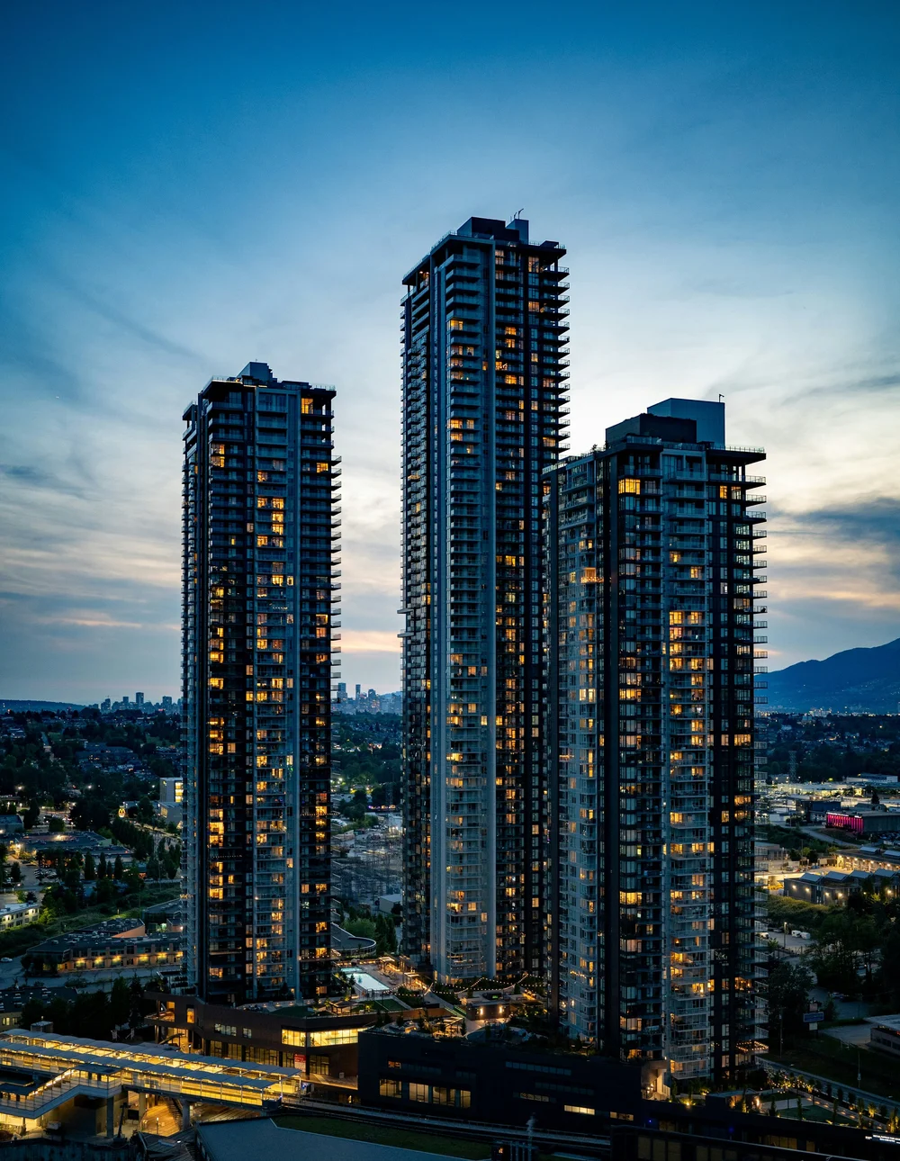 Three illuminated high-rise residential towers at dusk above a city skyline