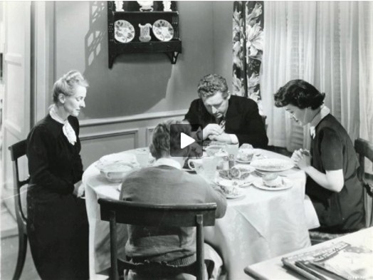A black and white photo showing people praying before eating at a table.