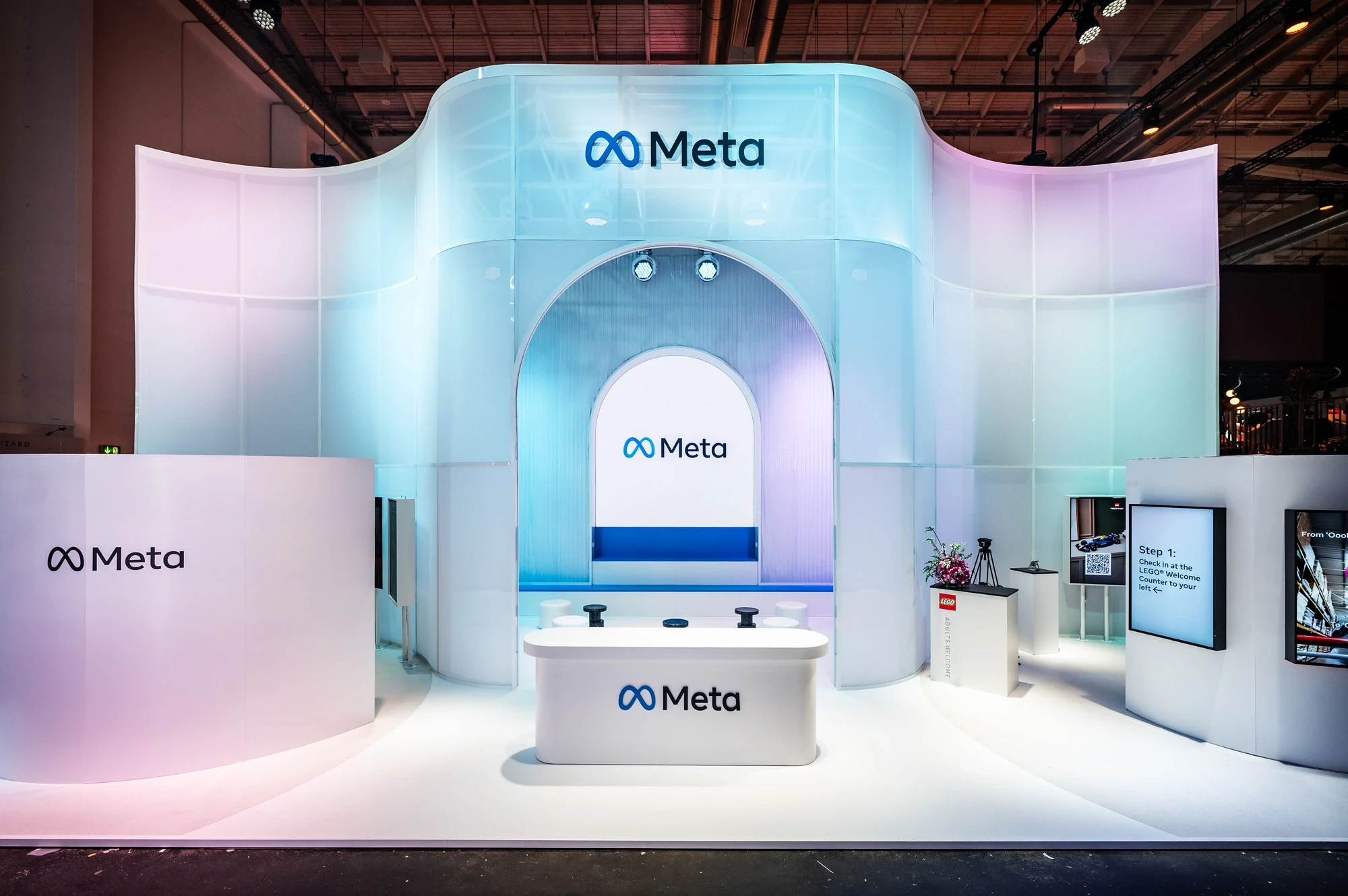 Meta exhibition booth featuring a sweeping arched architecture in white and soft blue light, central Meta logo above a sculpted reception counter.