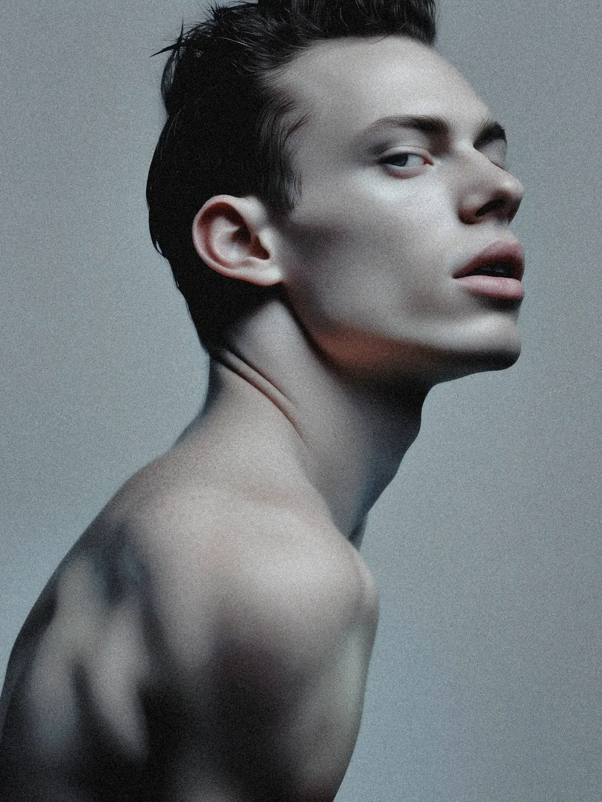 Close-up portrait in three-quarter profile with bare shoulders against a cool gray background.