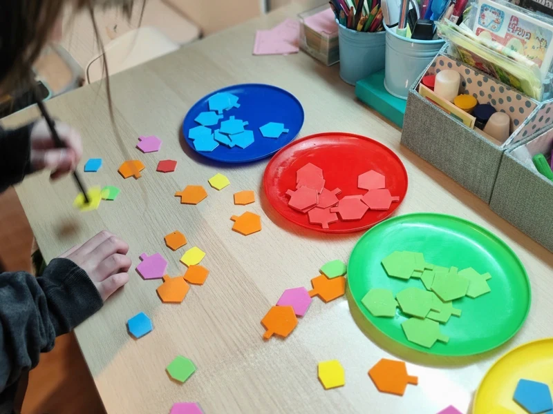 מבט מקרוב על פעילות יצירה וריפוי בעיסוק בקליניקה של 'נעים ולומדים': ילד עובד עם אלמנטים צבעוניים בצורת סביבונים וממיין אותם לפי צלחות צבעוניות. הפעילות מתבצעת על שולחן עץ באווירה לימודית ומפתחת.