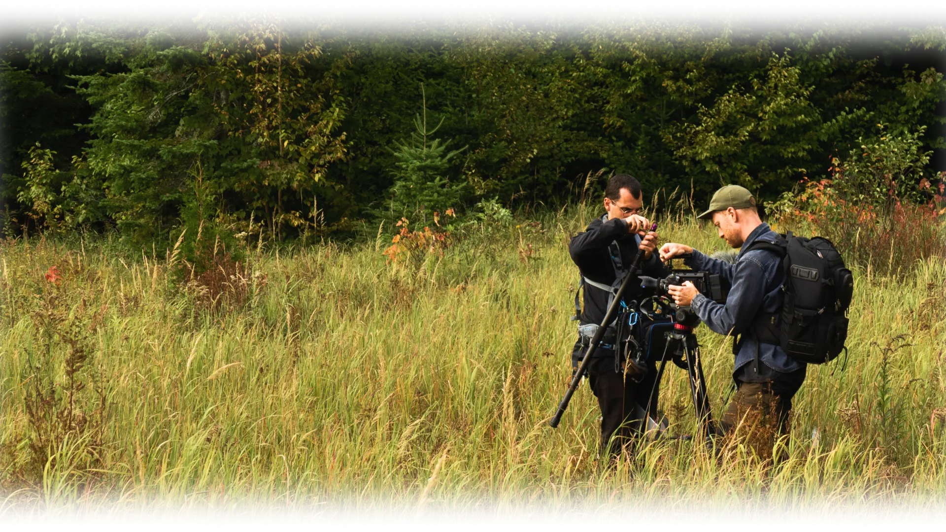 Deux personnes ajustant du matériel audio et vidéo sur un trépied au milieu d’une prairie, entourées d’herbes hautes et d’une forêt dense.