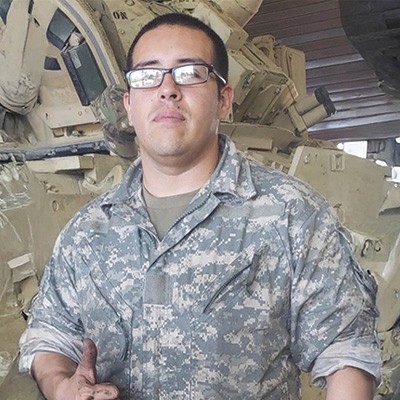 A soldier wearing fatigues shown standing before heavy equipment and giving a "thumbs up" gesture