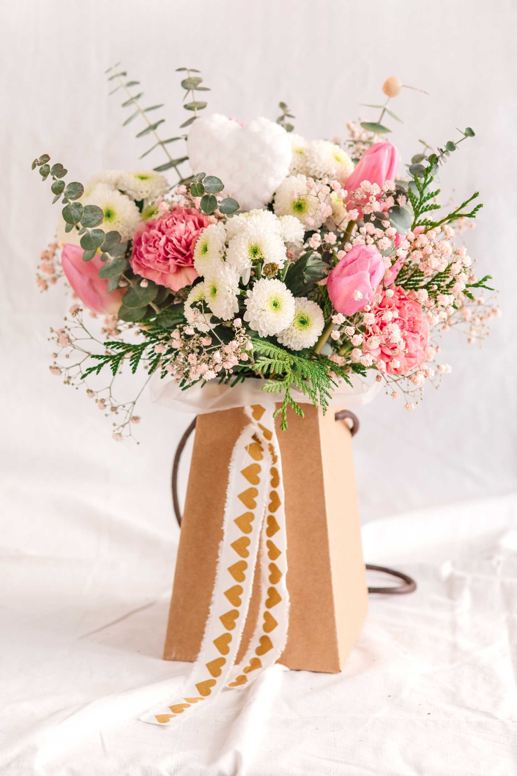Arranjament floral en tons rosa i blanc amb tulipes, clavells i margarides dins una bossa de cartró kraft, decorada amb cinta blanca amb cors grocs.