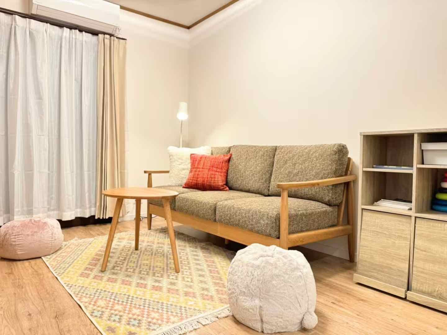 心地よいリビングルームには、プラッシュなベージュソファに赤いアクセントピローがあり、丸い木製のコーヒーテーブル、パターン柄のラグに置かれた柔らかいプーフ、装飾品が飾られた木製の本棚が備えられています。これらのすべてが、軽いカーテン付きの大きな窓から差し込む自然光で暖かく照らされています。この空間は民泊運営において資産運用の安定性を高め、京都の専門知識を生かして収益性を高めることを目指しています。