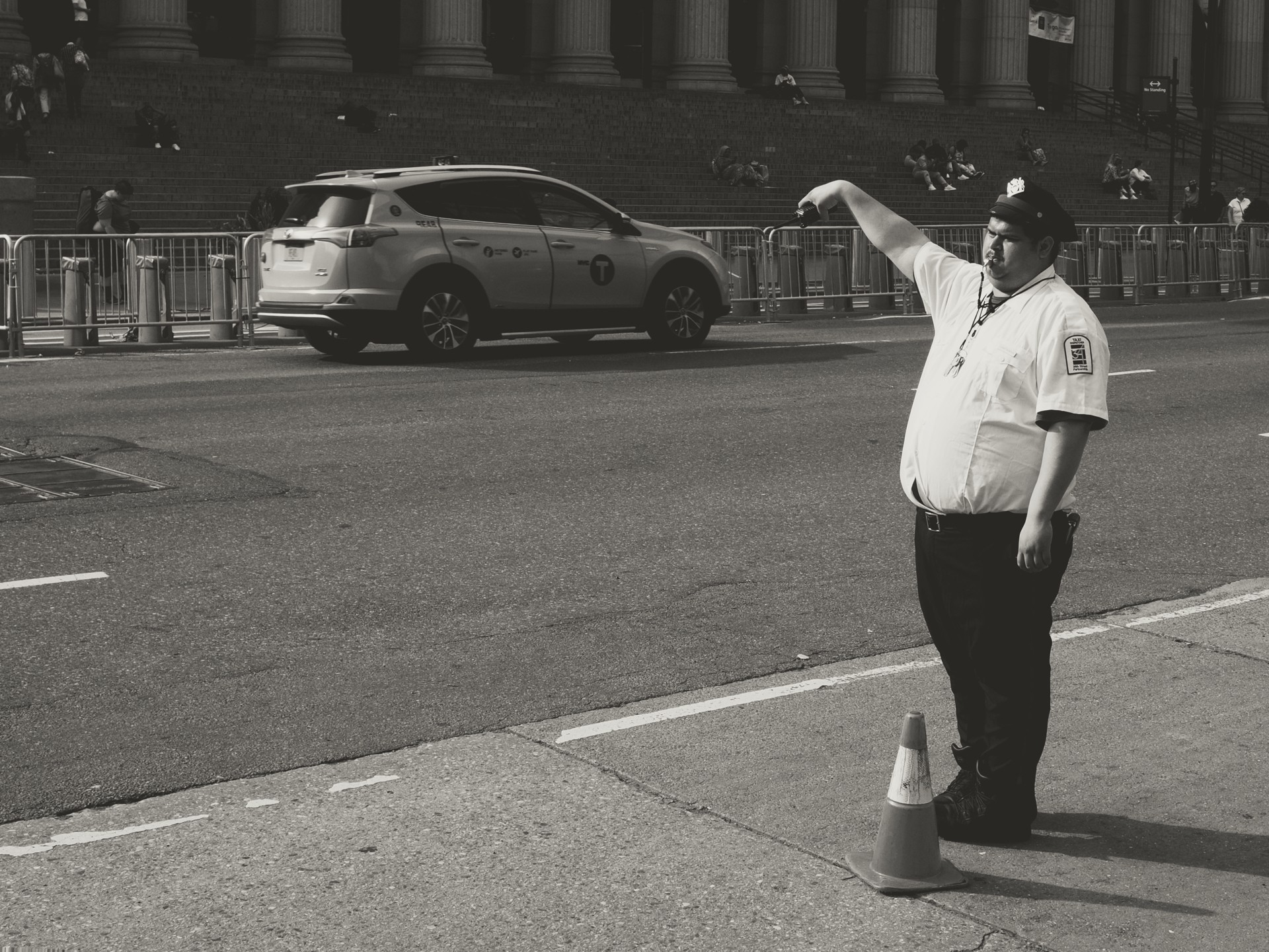 Black-and-white street scene showing a uniformed traffic officer directing traffic beside an orange cone, a white SUV taxi passing, metal barricades, and a grand building with tall columns and seated pedestrians on the steps.