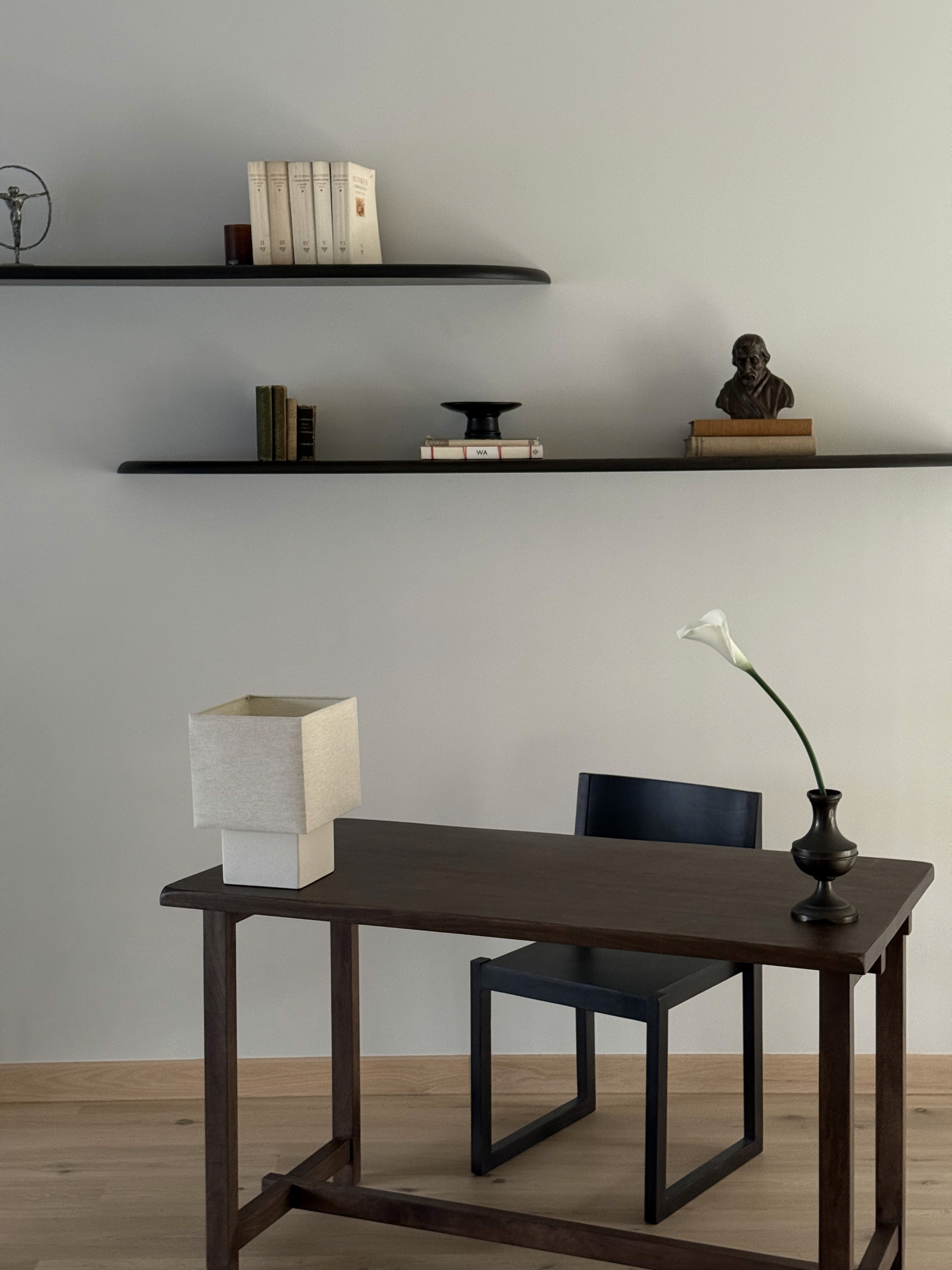 Minimalist home office with dark walnut writing desk, black stool, linen table lamp, and floating shelves with books and bronze bust sculpture