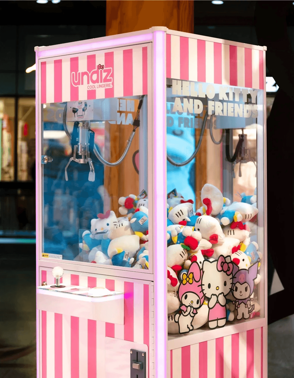 Machine à pince rose et blanche remplie de peluches Hello Kitty, avec le logo "undiz" et "HELLO KITTY AND FRIENDS".