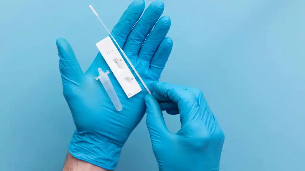 Blue-gloved hands holding a white lateral flow test cassette and a sample tube against a blue background
