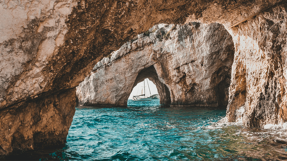 A serene view of a yacht sailing through turquoise waters beneath the natural arches of a rocky coastal cave, perfect for a peaceful yachting retreat and yoga escape.