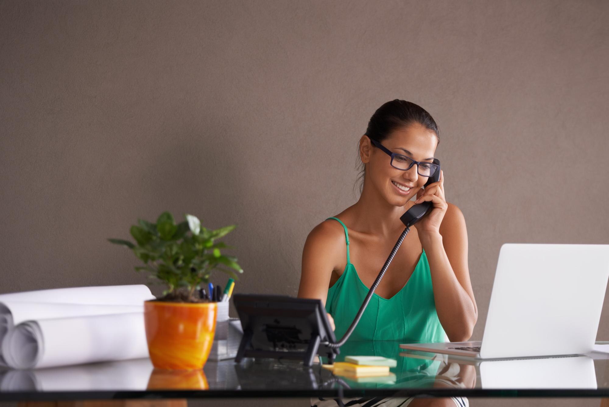 Vrouw met bril beantwoordt een vaste telefoon op kantoor met laptop