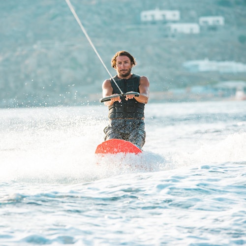 Person wakeboarding on water, wearing a life vest and holding onto a tow rope, with a mountainous background and a few buildings.
