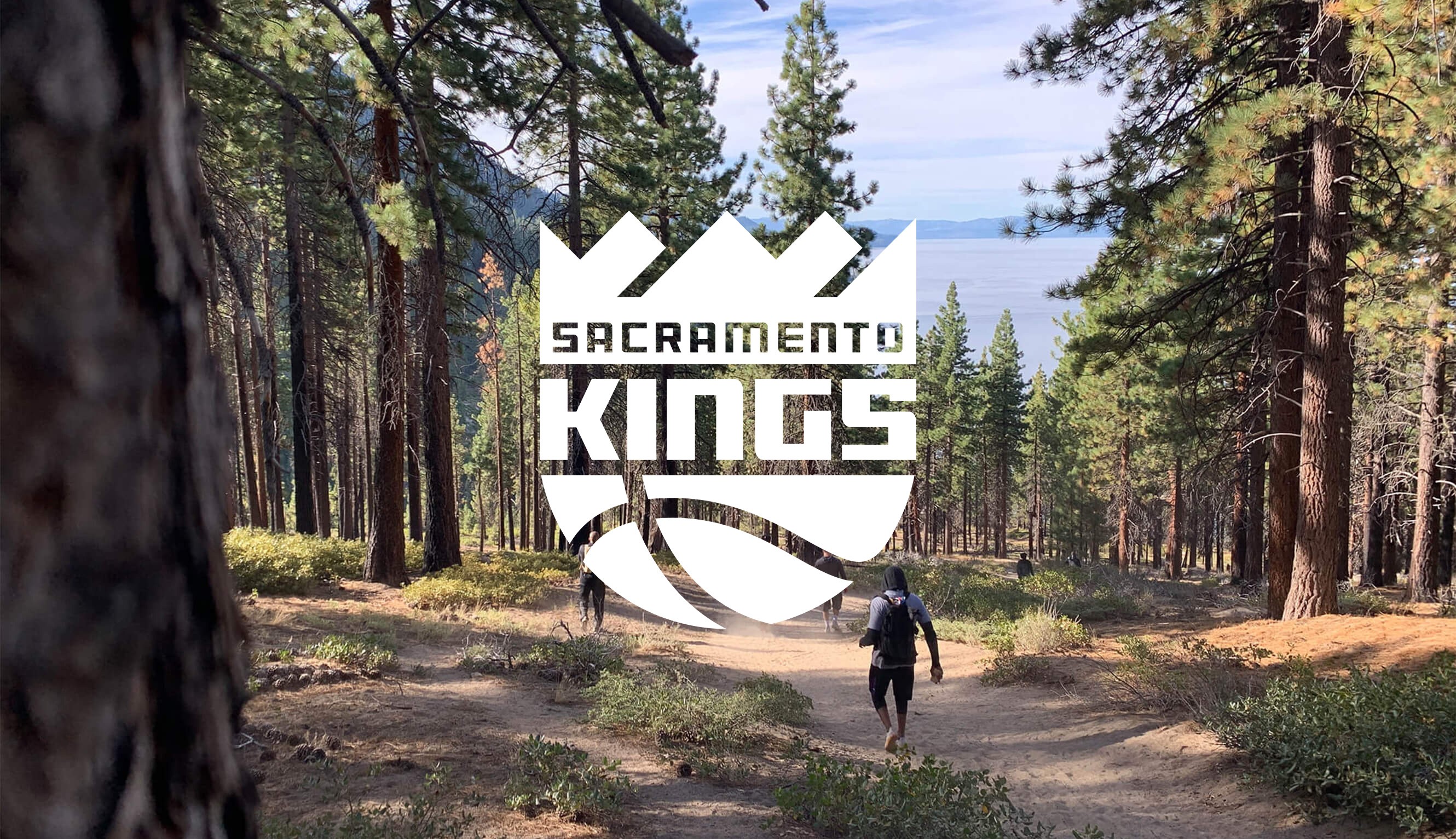 A forest, with poeple walking down a trail and the Sacramento Kings logo in white on top of the image