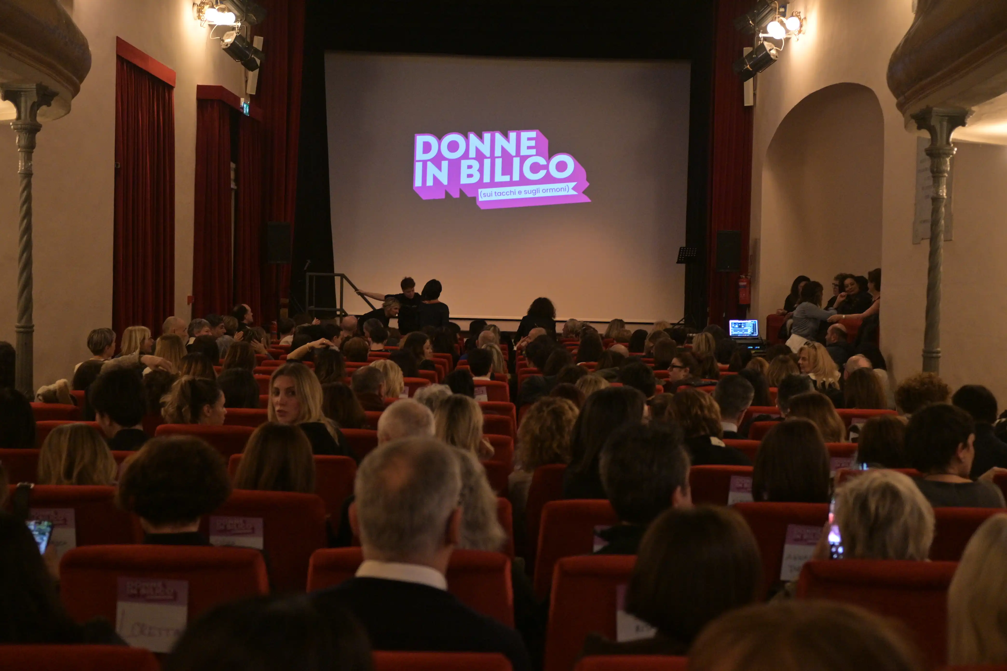 Pubblico seduto in sala al teatro verdi di Forlimpopoli con logo Donne in Bilico proiettato a schermo