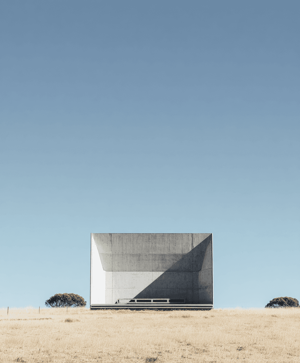 A minimalist concrete pavilion sits alone on a dry, golden field beneath an expansive clear-blue sky, evoking calm isolation.