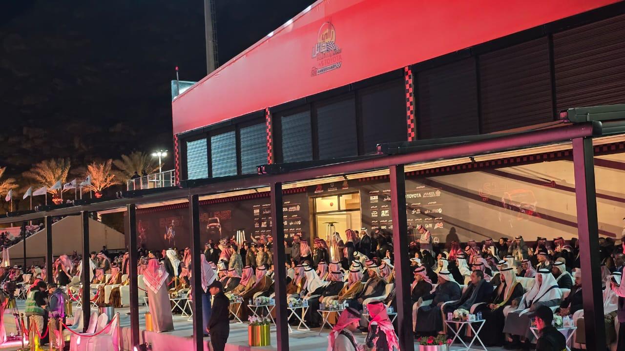 Audience seated in an outdoor venue, many wearing traditional attire, beneath a covered area at night.