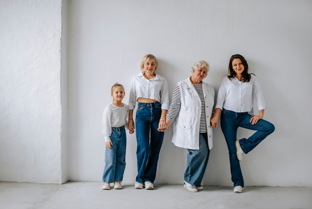 A multi-generational family portrait featuring four women from different age groups, all dressed in casual white tops and blue jeans. They stand against a plain white wall, exuding a sense of unity and familial bond, celebrating the connections and heritage shared across generations.