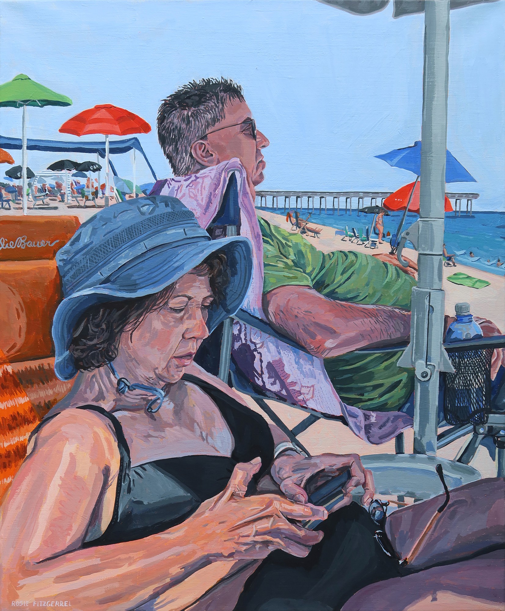 Two people sit in bathing suits on the beach, under the shade of the umbrella. One is looking at a phone, while the other looks out to the ocean.