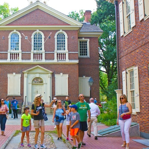 一群人，包括孩子们，正在带有拱形窗户和白色百叶窗的历史砖建筑附近漫步和探索。