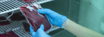 Gloved hands holding a sealed bag of blood inside a medical storage unit.