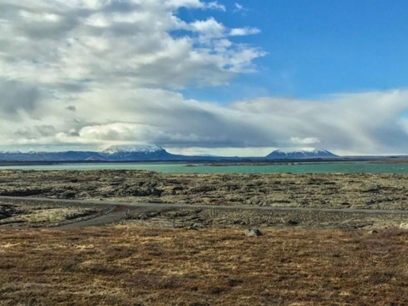 Wide view across the lava-covered shores of Lake Mývatn in North Iceland, with large volcanic craters and flat-topped tuya mountains rising in the distance beneath dramatic clouds and blue sky.