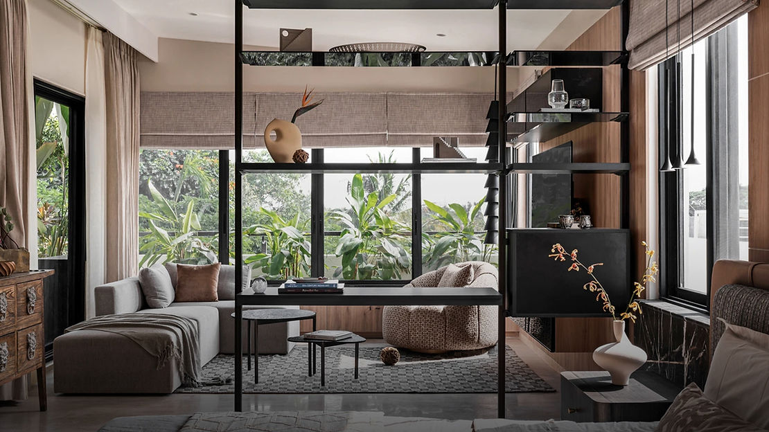A wide, open living room with big windows and green trees outside. A black shelf divides the room at the halfway point. A big grey rug covers most of the floor. Grey and brown sofas are on the side of the room, closer to the windows. The black shelf has ornamental jugs and books.