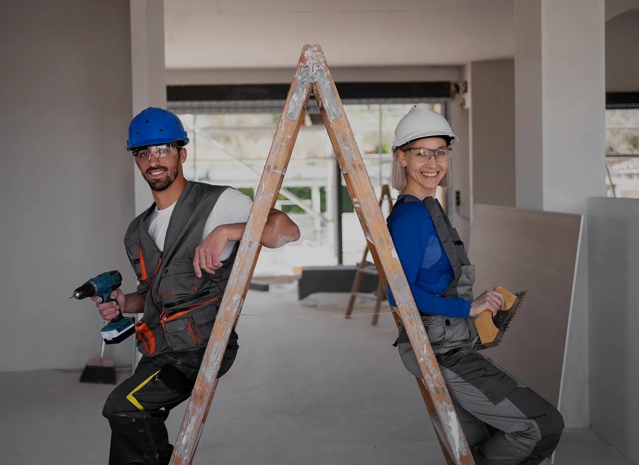 Two construction workers, a man and a woman, sit back-to-back on a ladder in a bright room, holding tools, wearing safety helmets and smiling confidently.
