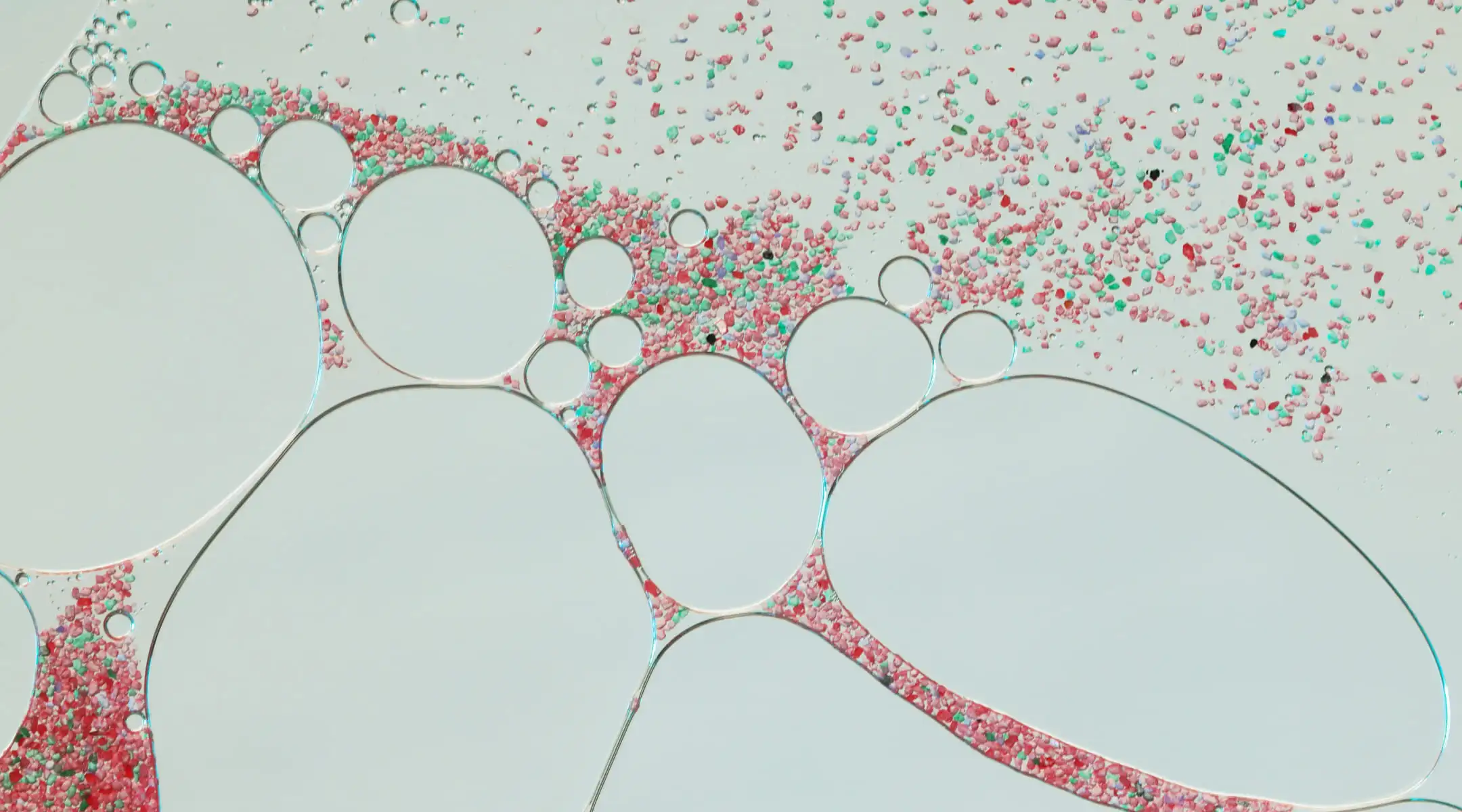 Abstract macro shot of clear liquid bubbles on a light grey background with small pink and green particles.