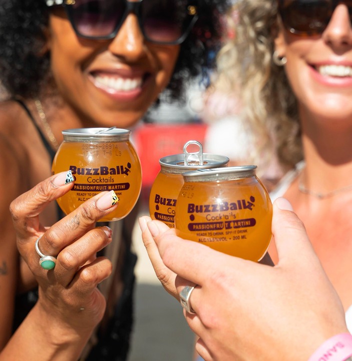 Three people holding chilled 200ml BuzzBallz Passionfruit Martini cocktails at a party. These bar-strength, ready-to-drink BuzzBallz are available for fast delivery on Gopuff UK along with 1.75L Biggies.
