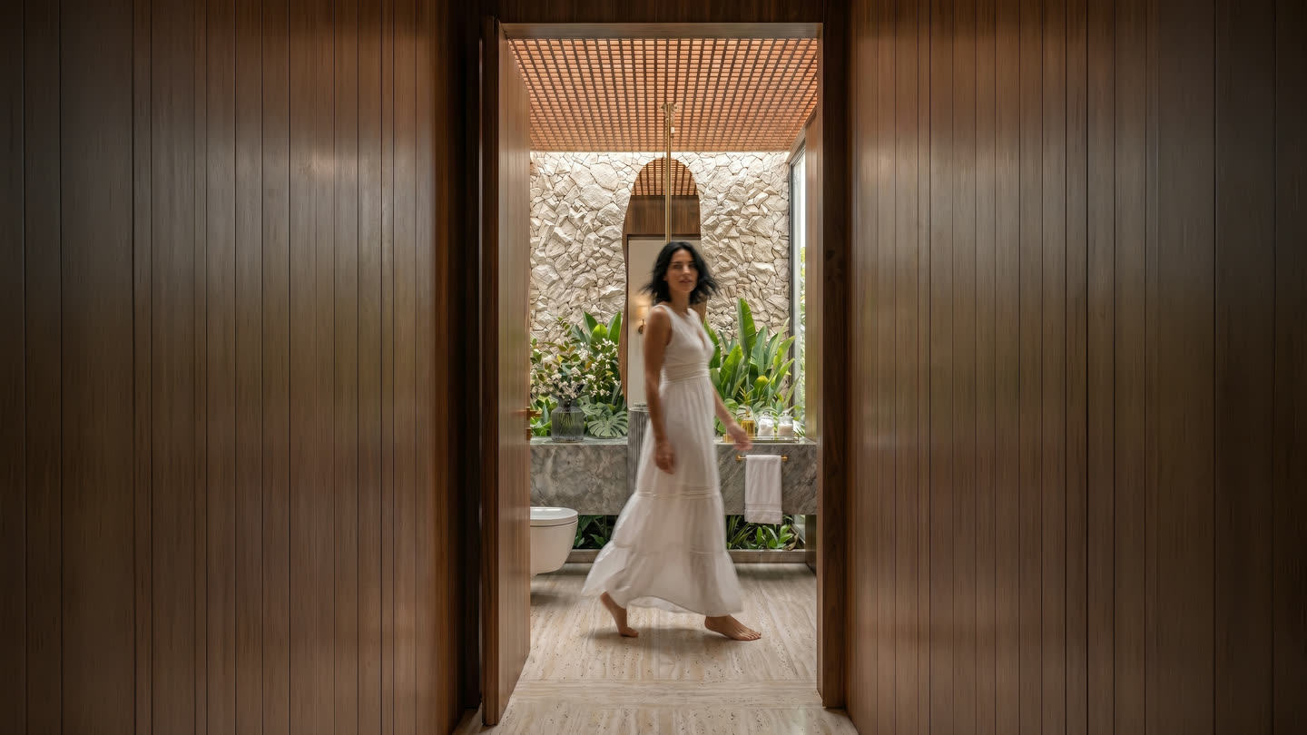 Mulher de vestido branco atravessando o corredor de madeira de uma suíte, com o movimento levemente desfocado. Ao fundo, o banheiro com parede de pedra e vegetação.