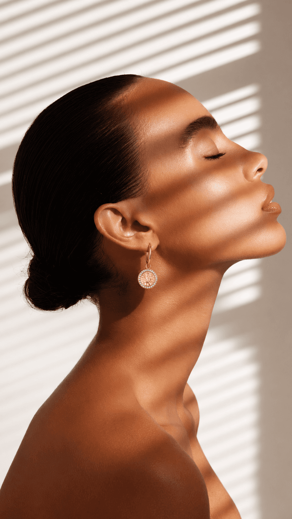 Profile of woman with closed eyes, smooth skin, and an golden earring, with light and shadow from blinds on her face.
