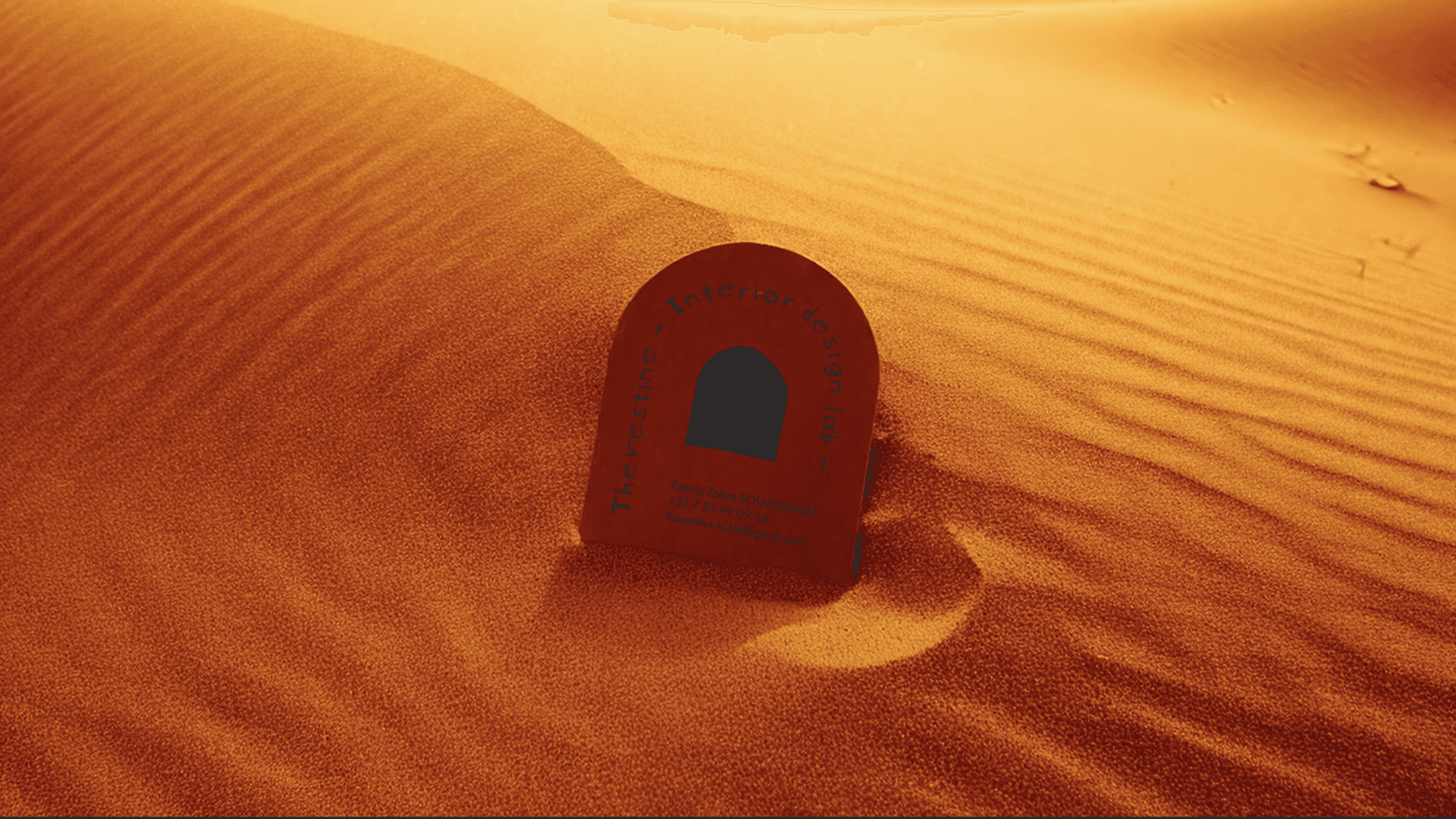 Une carte de visite de "THEVESTINE" enfouie partiellement dans le sable, avec un design épuré et un logo minimaliste. Elle reflète l'approche avant-gardiste et ancrée dans la nature de "THEVESTINE", fusionnant modernité et traditions architecturales.