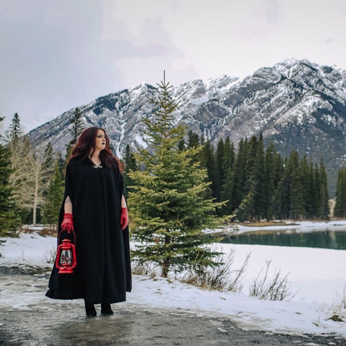 A woman in a black cloak holding a red lantern stands by a snowy lake with evergreen trees and mountains in the background.