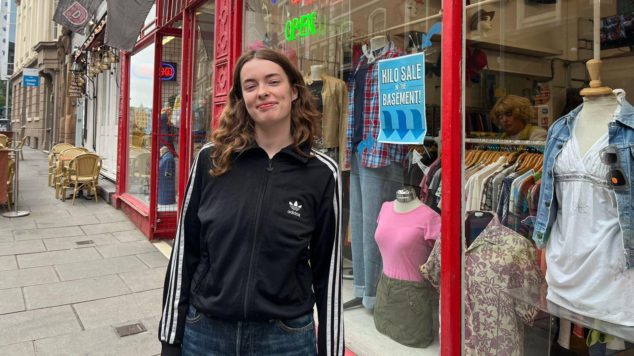 A woman wearing a black zip up Adidas jacket. She has dark brown curly hair. In the background is her shop which has a red front and 