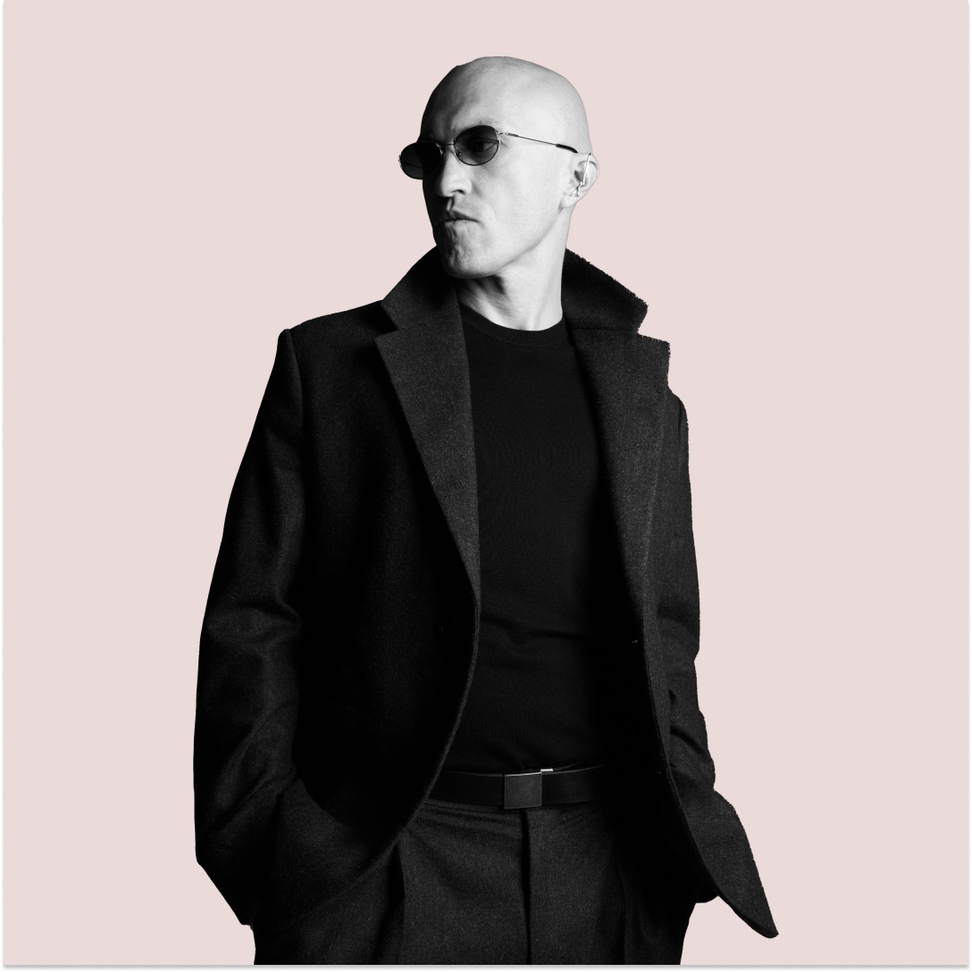 Casual studio portrait of a bearded man with curly hair in a white shirt, arms crossed, on a light gray background.