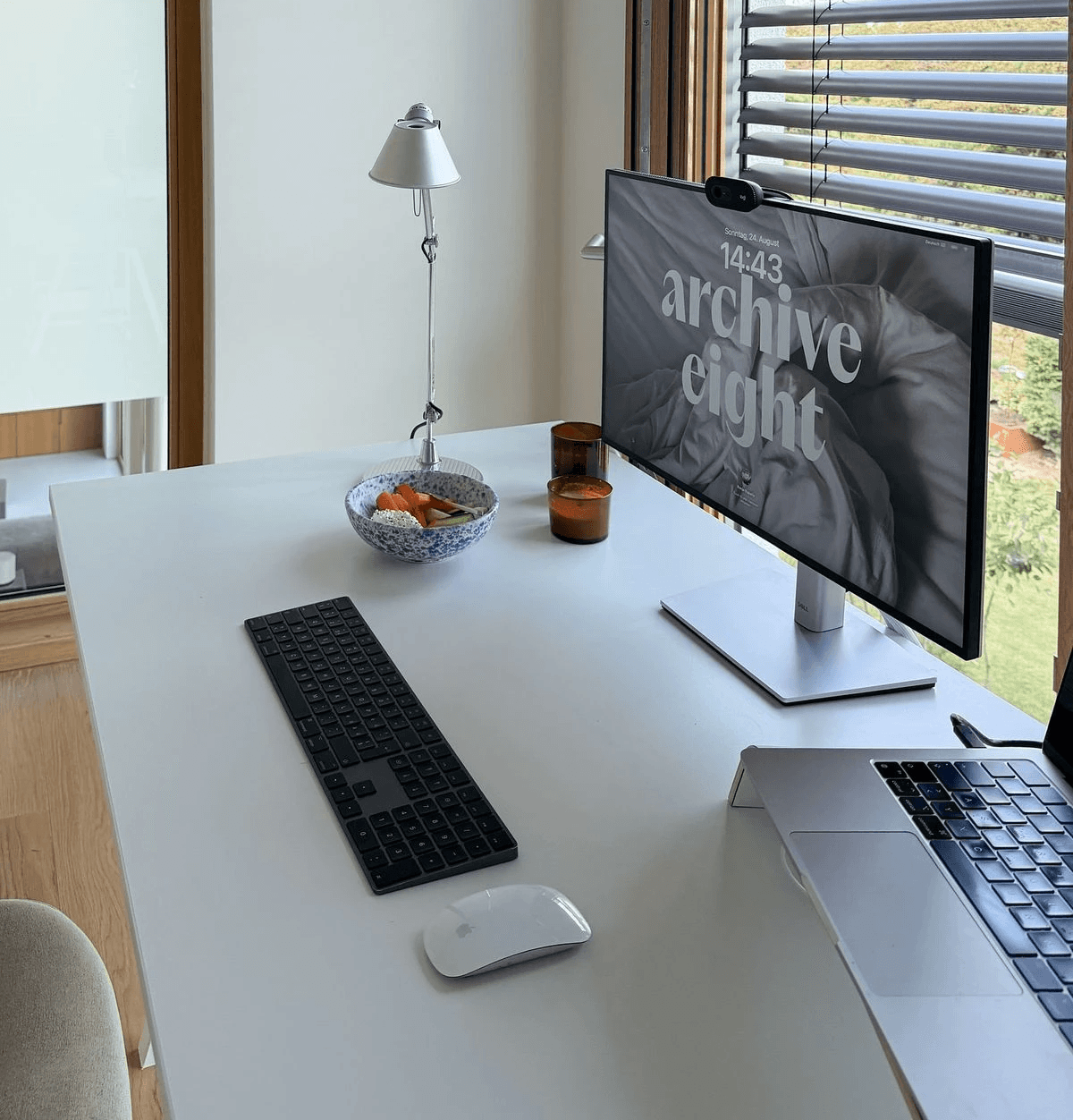 Modern home office setup with a monitor, ergonomic chair, keyboard, and indoor plants near a window with blinds.