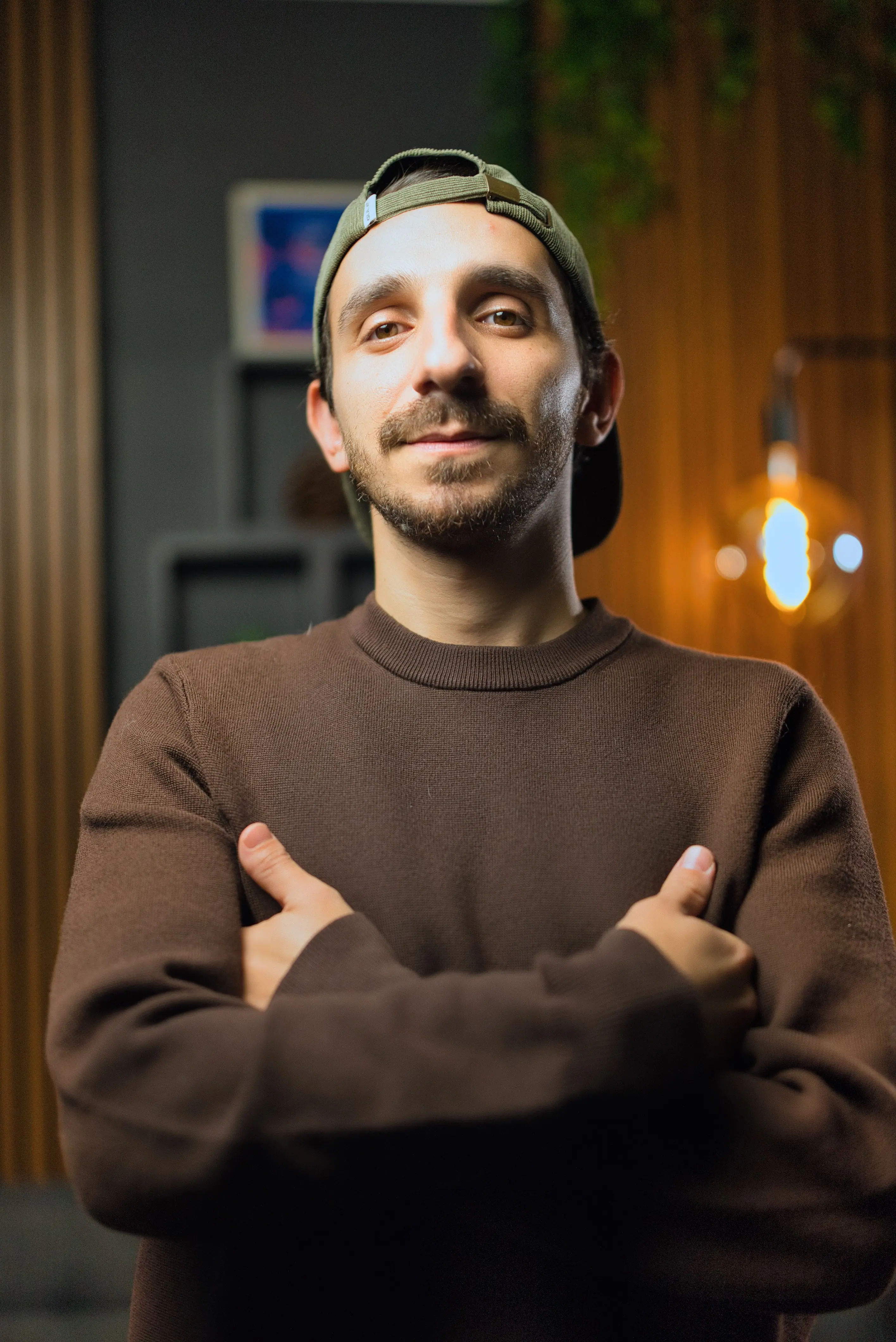 A man wearing a brown sweatshirt and a backwards cap stands confidently with arms crossed in a warmly-lit room featuring wooden paneling and an industrial-style light fixture.