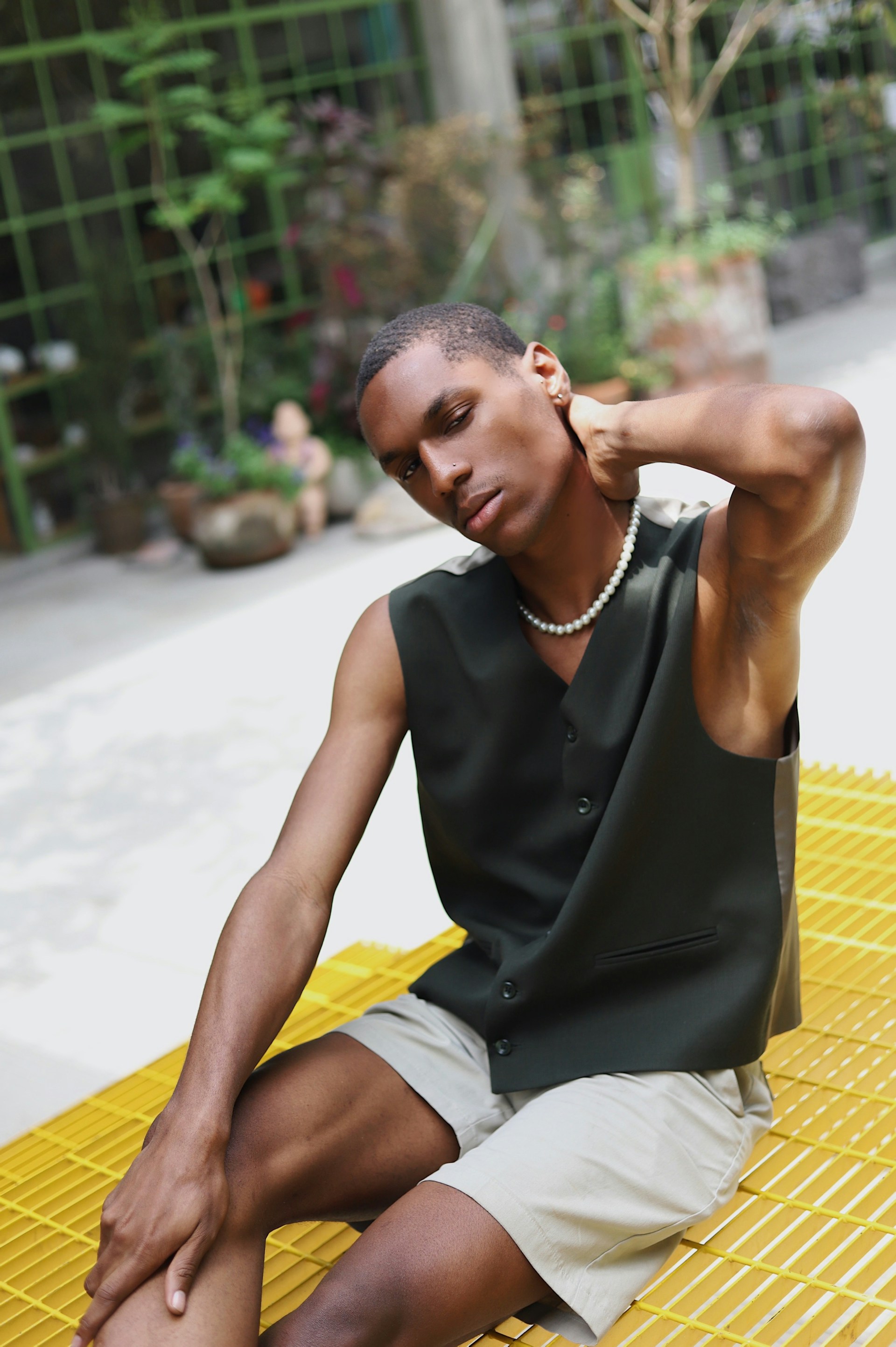 A close-up of a man wearing a pearl necklace and dark green vest, sitting on a yellow metal grate bench. He has one hand behind his neck and looks toward the camera with a neutral expression.