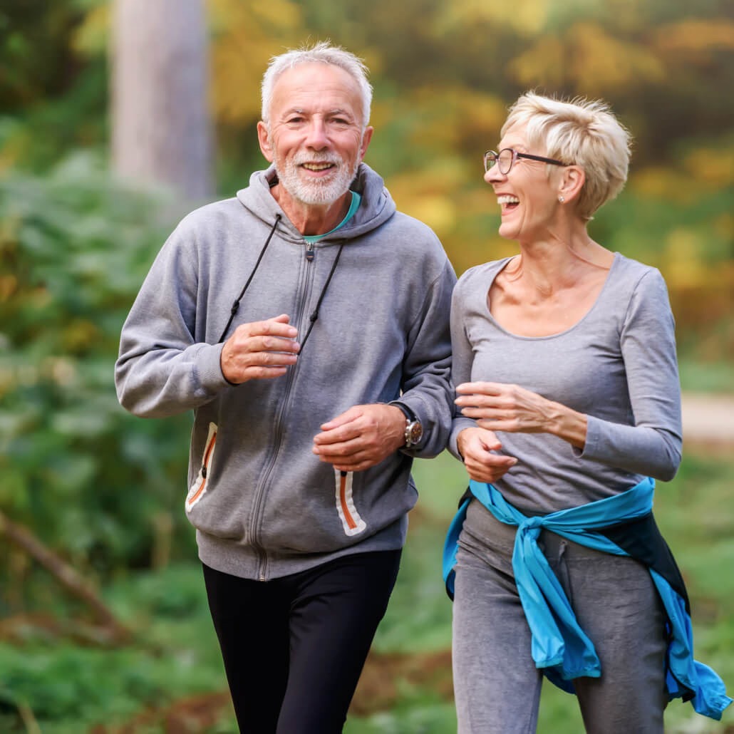 An older couple in athletic wear jog outdoors, laughing.