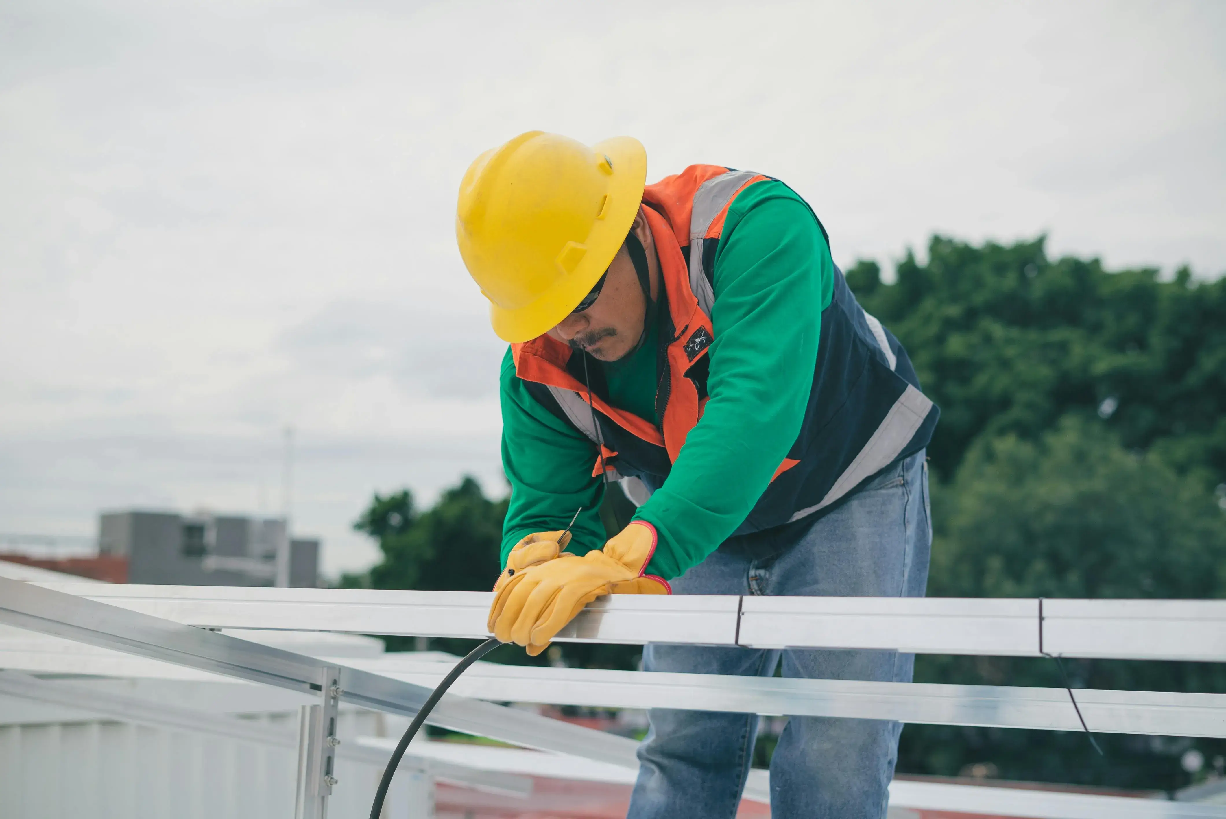 A construction worker in a yellow hard hat and safety vest is installing metal beams on a rooftop. The background features cloudy skies and greenery.