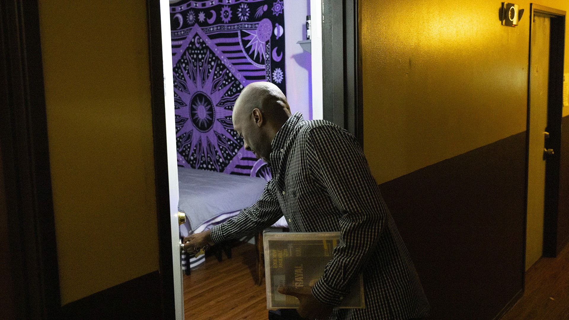 Darnell Price leans over and uses his right hand to close a door into a room with a bed and a printed fabric displayed on a wall while standing in a hallway with emergency lights and another door.