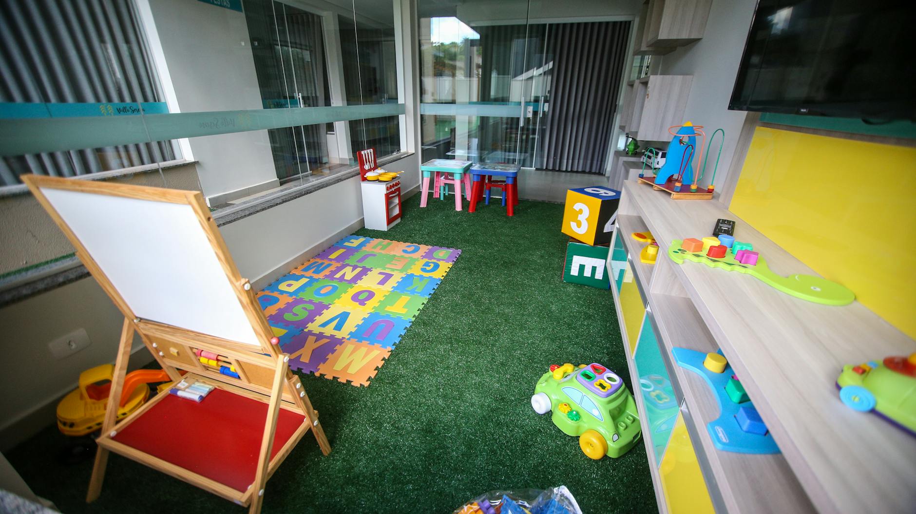 A smiling kindergarten teacher points to a colorful alphabet chart while young students raise their hands to participate.