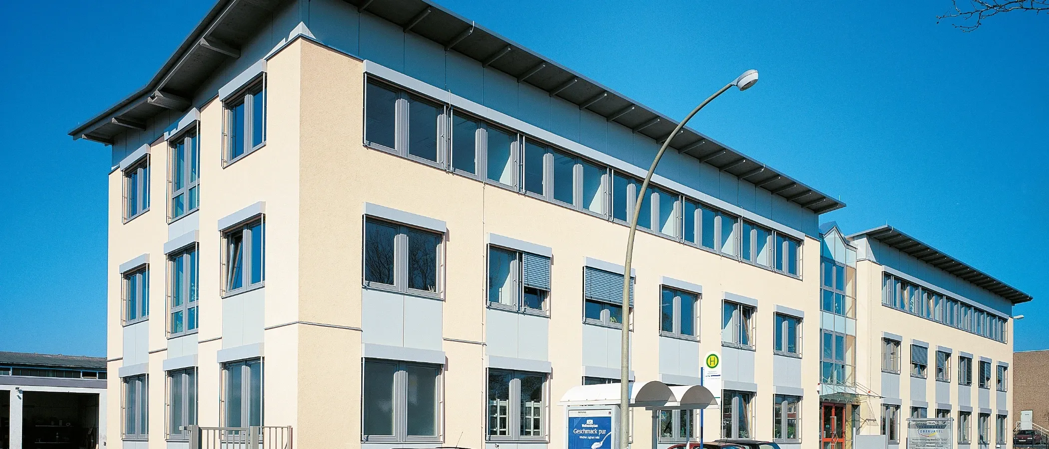 Moderne Bürofassade in hellem Gelb mit grau abgesetzten Fensterrahmen und Dachvorsprüngen, an einem klaren blauen Himmel.