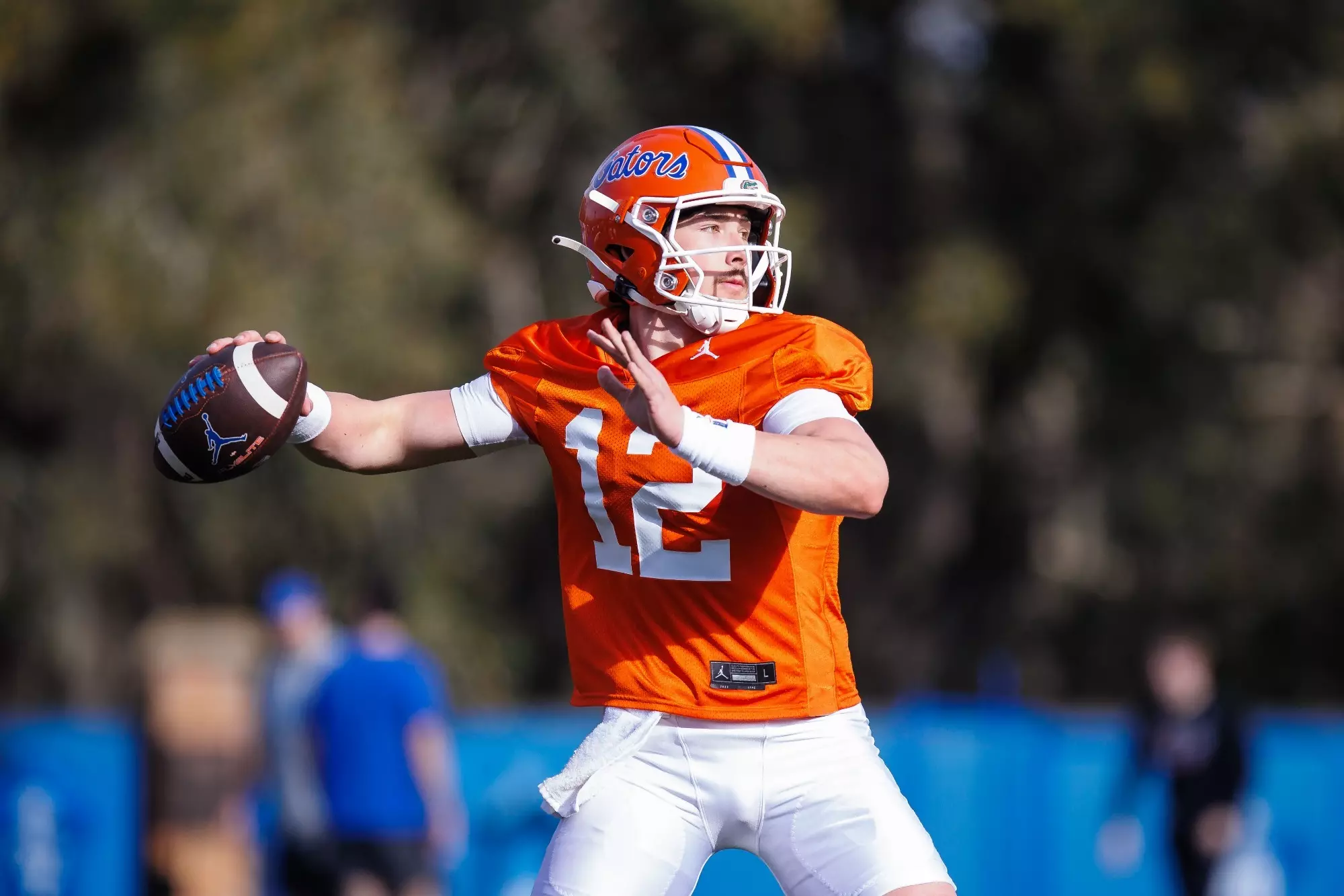 Aaron Philo in an orange jersey and helmet prepares to throw a football during practice. He stands outdoors with trees and blurred figures in the background.