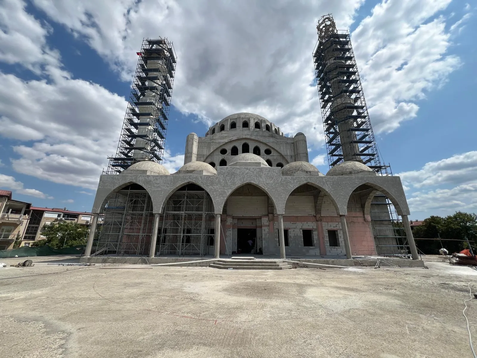 Front view of a mosque under construction in Skopje, featuring domed arches, two minarets surrounded by scaffolding, and ongoing structural works. Supervision by Terraplan Partners.