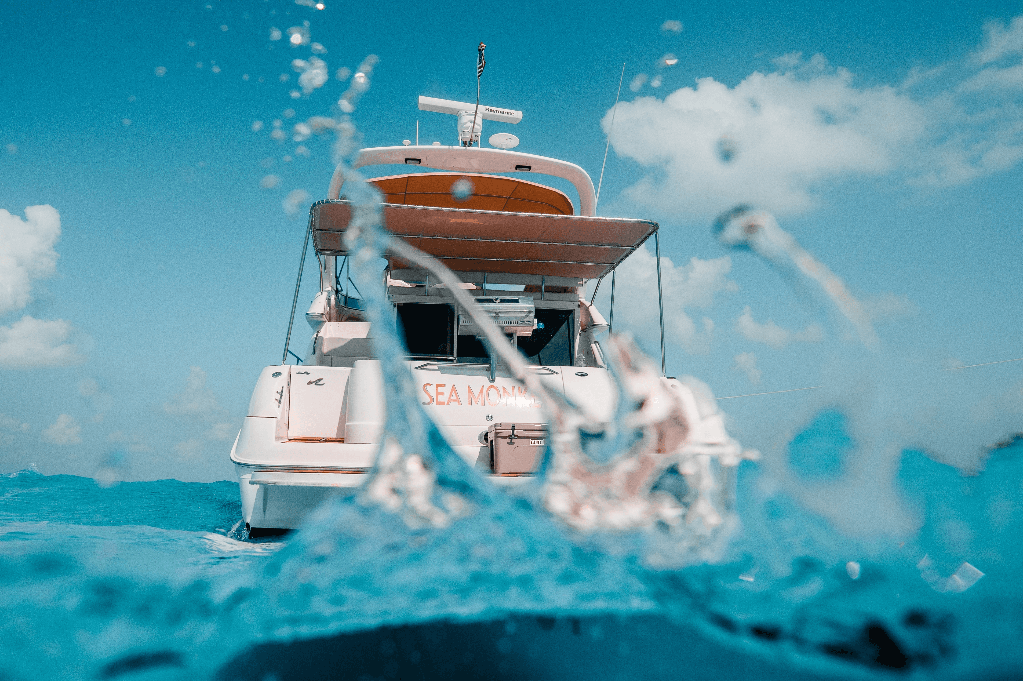 Luxury yacht viewed from water level with ocean splash in foreground beneath a bright blue sky.