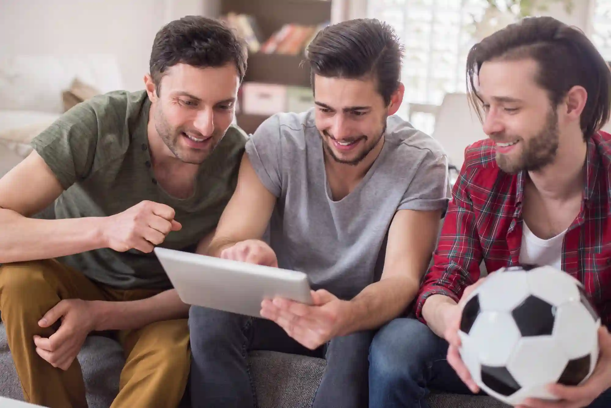 Three male friends sit together on a couch, holding a soccer ball while looking at a digital tablet.