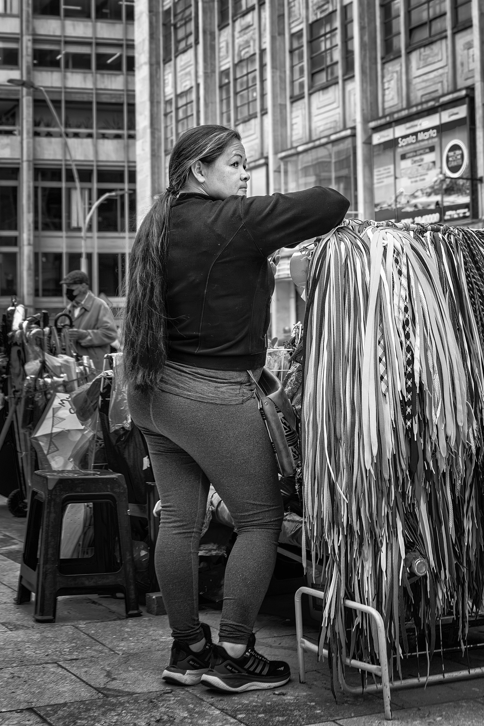 A woman stands beside a rack of hanging shoelaces or ribbons at a bustling Bogotá street market, glancing back over her shoulder.